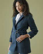 A woman with curly dark hair models a dark teal belted blazer over a white ruffled blouse and plaid trousers, set against a solid olive green studio background.
