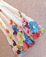 A sheer white scarf with a vibrant multicolor floral print, laid out diagonally on a textured blush herringbone fabric background.