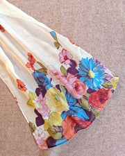 A sheer white scarf with a vibrant multicolor floral print, laid out diagonally on a textured blush herringbone fabric background.